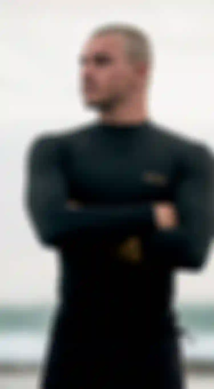 Man in black wetsuit with arms crossed stands on a beach, looking sideways. Waves and a cloudy sky in the background.