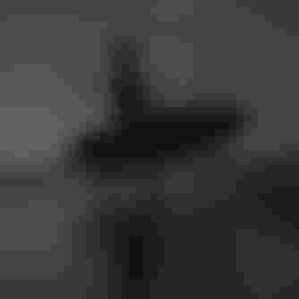 Silhouetted person walking on a beach, carrying a windsurfing board, with a cloudy sky and distant hills in the background.