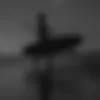 Silhouetted person walking on a beach, carrying a windsurfing board, with a cloudy sky and distant hills in the background.