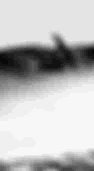 Silhouetted kiteboarder mid-air against a bright sky, with water spray and distant hills in the background.