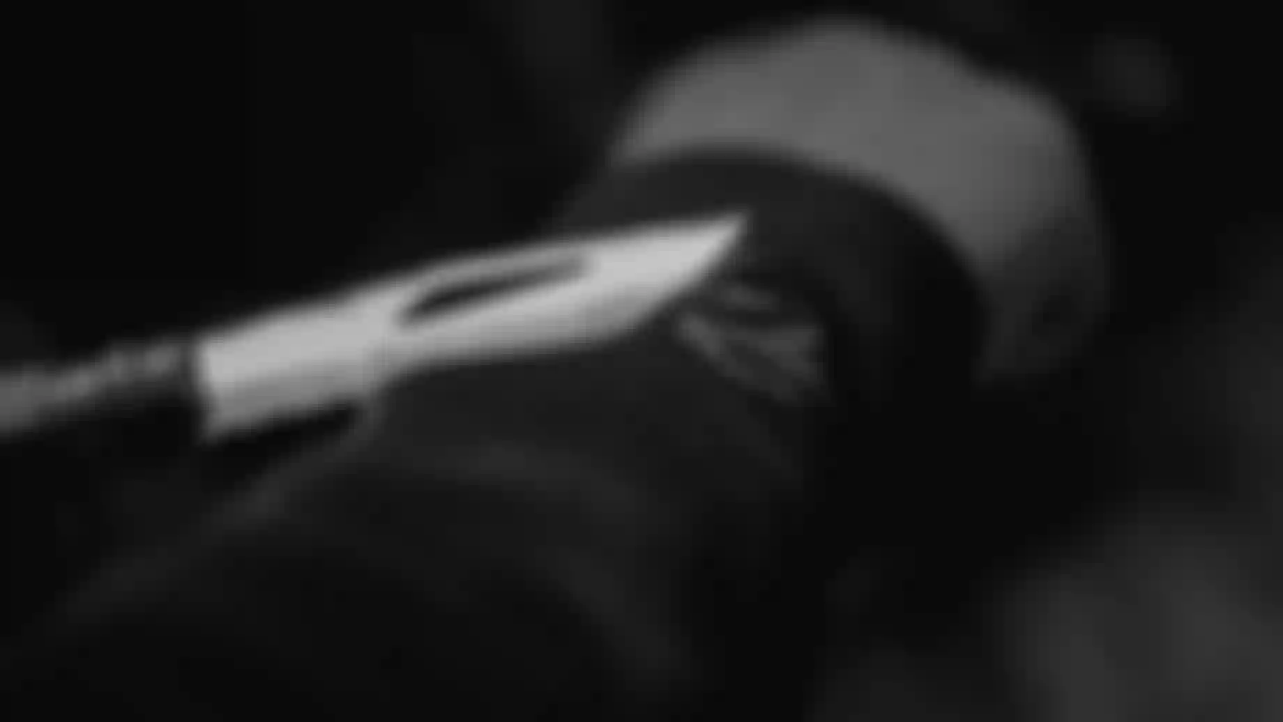 Close-up of a person holding a knife to their arm, with the blade touching a dark fabric sleeve. The image is in black and white.