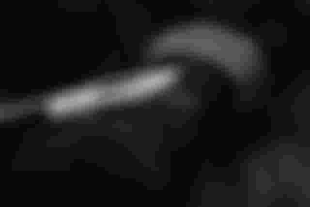 Close-up of a person holding a knife to their arm, with the blade touching a dark fabric sleeve. The image is in black and white.