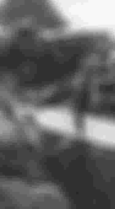 A person holding a surfboard walks over rocky terrain by the ocean, with waves crashing in the background. Black and white image.