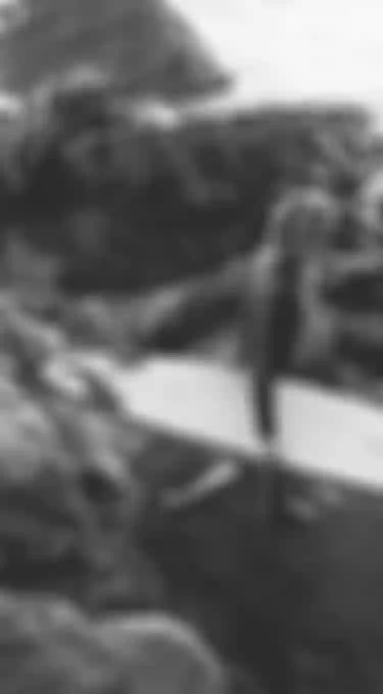 A person holding a surfboard walks over rocky terrain by the ocean, with waves crashing in the background. Black and white image.