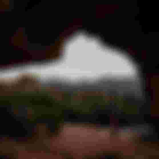 Person standing in a cave opening looking out over red-rock desert, green shrubs and distant mesas at dusk.