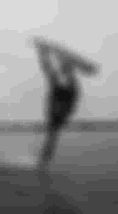 Person kiteboarding, performing an aerial trick over water, creating a splash under a cloudy sky. Black and white tone.