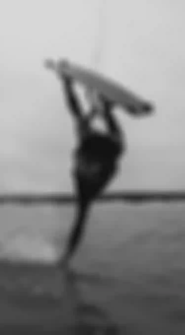 Person kiteboarding, performing an aerial trick over water, creating a splash under a cloudy sky. Black and white tone.
