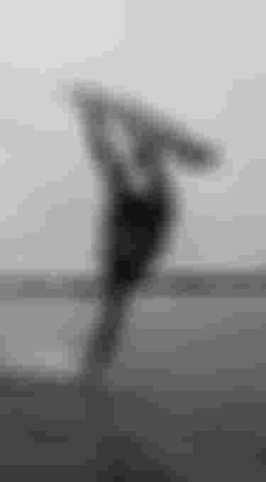 A kiteboarder performs an acrobatic trick over the water, lifting the board high, with a backdrop of a cloudy sky and distant shoreline.