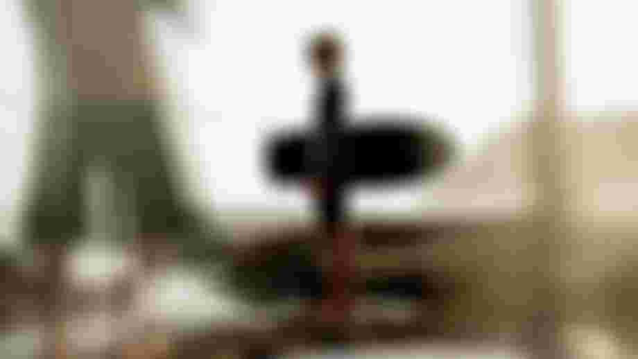 A surfer in a wetsuit stands on rocky shore holding a board, gazing at the ocean with cliffs in the background.