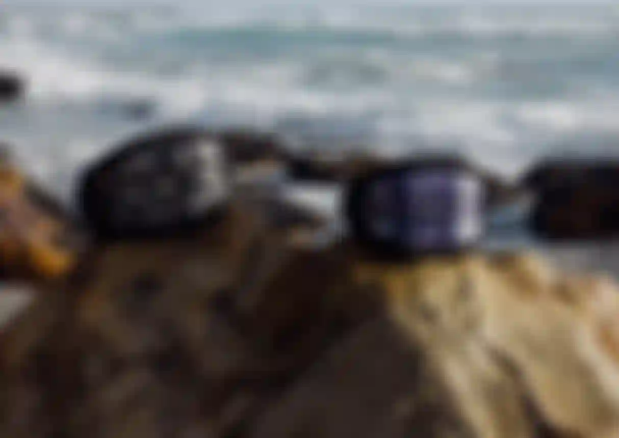 Two waist packs are placed on rocks by the ocean, with waves in the background. One is black, the other has a purple pattern.