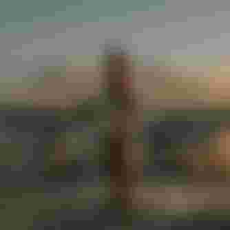 A person in a wetsuit stands on a beach holding a kiteboard, with kites and distant mountains in the background during sunset.