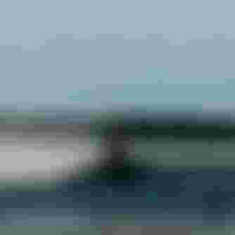 A person kiteboarding over the water, creating a spray. They are leaning back, holding the kite's control bar, with a rocky shoreline in the background.