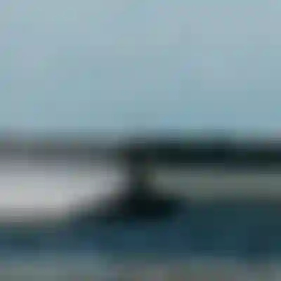 A person kiteboarding over the water, creating a spray. They are leaning back, holding the kite's control bar, with a rocky shoreline in the background.