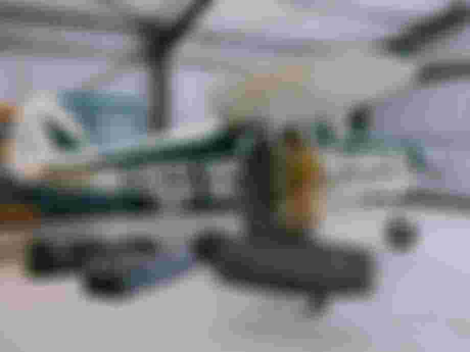 A person carries a surfboard near a vintage green and white airplane in a hangar, with multiple bags on the floor.