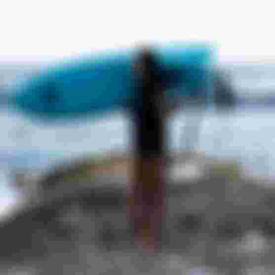 ION athlete Mo Chauvin standing on the coast in Guadeloupe with a pronefoil on her shoulder, wearing an ION Amaze V-Back wetsuit.
