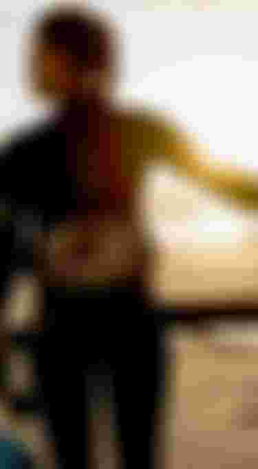 A woman in a wetsuit stands on a beach at sunset, holding a surfboard and sail, looking towards the sea.