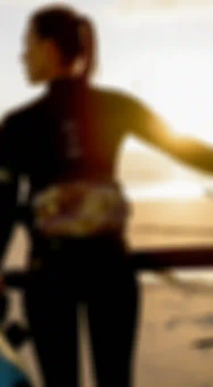 A woman in a wetsuit stands on a beach at sunset, holding a surfboard and sail, looking towards the sea.