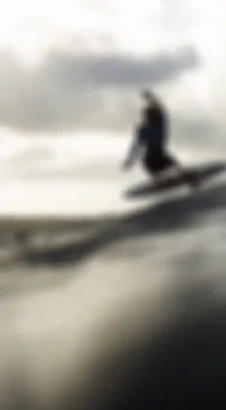 Surfer catching air above a wave, with cloudy sky and distant shoreline in the background.