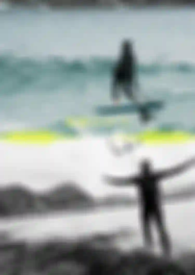 Two wetsuit-clad riders: one hydrofoil surfing ocean waves, one celebrating on a rocky shoreline with arms outstretched and kite bar overhead.