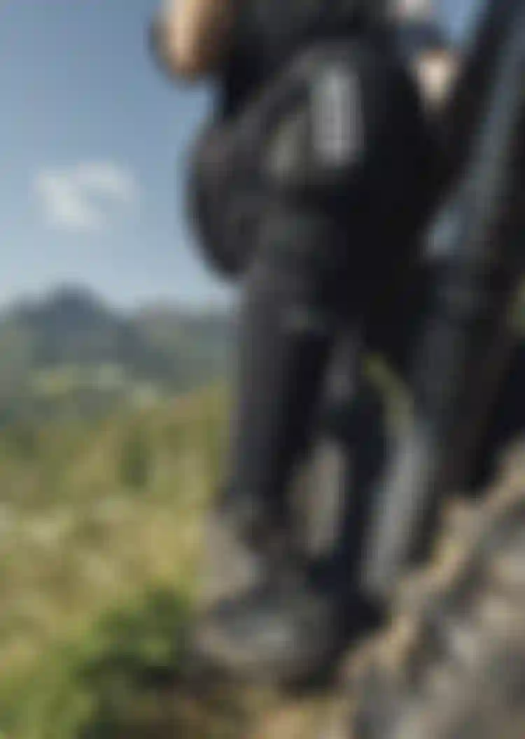 Close-up of a mountain biker's legs and bike on a grassy hill, with protective gear and rugged mountains in the background under a blue sky.