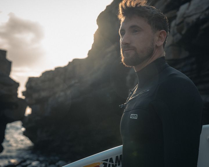 Man in a wetsuit with a surfboard stands by rocky cliffs at sunset, gazing towards the ocean.
