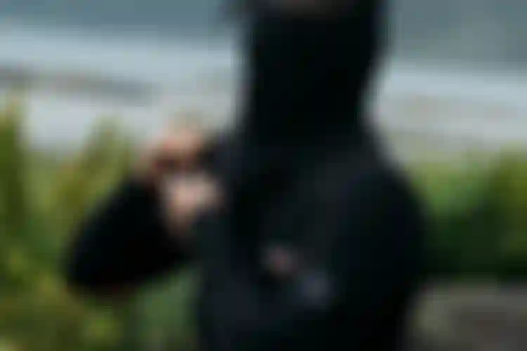 Person zipping up a black hooded wetsuit near a misty coastal landscape with mountains and water in the background.