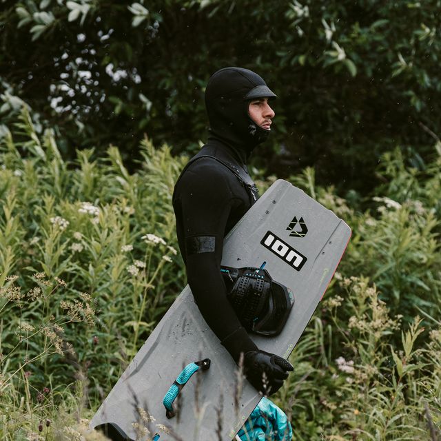 A person in a wetsuit and hood carries a kiteboard while walking through tall grass, with lush green foliage in the background.