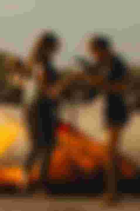 Two women on a sandy beach prepare kiteboarding gear, including a bright orange kite. One woman holds a harness, while the other wears sunglasses.