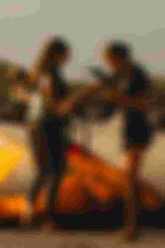 Two women on a sandy beach prepare kiteboarding gear, including a bright orange kite. One woman holds a harness, while the other wears sunglasses.