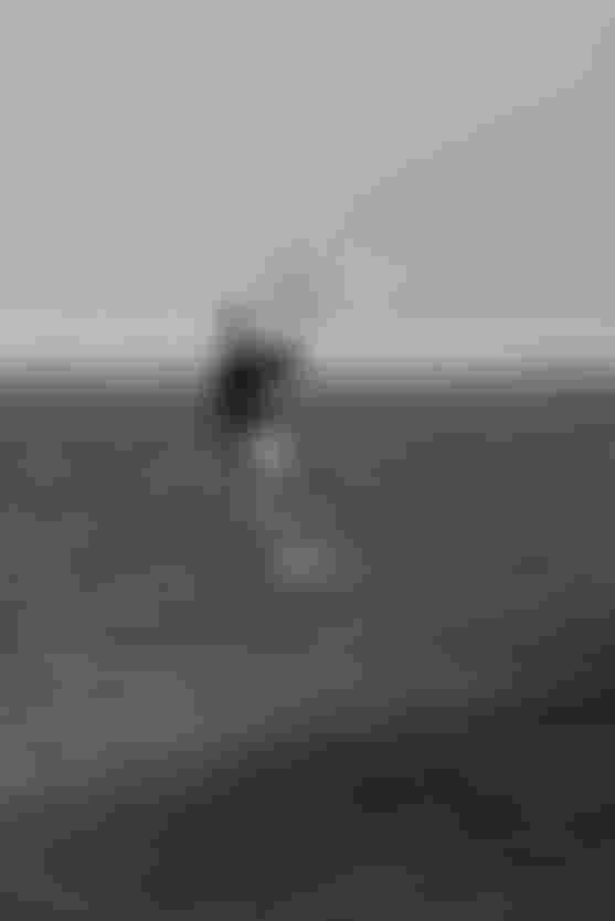 Person kitesurfing on a hydrofoil board in open water, wearing a wetsuit. The background features a distant, hazy shoreline.