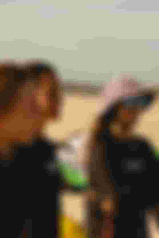 Three women wearing black shirts and one with a purple hat stand on a sunny beach, smiling and looking towards something in the distance.