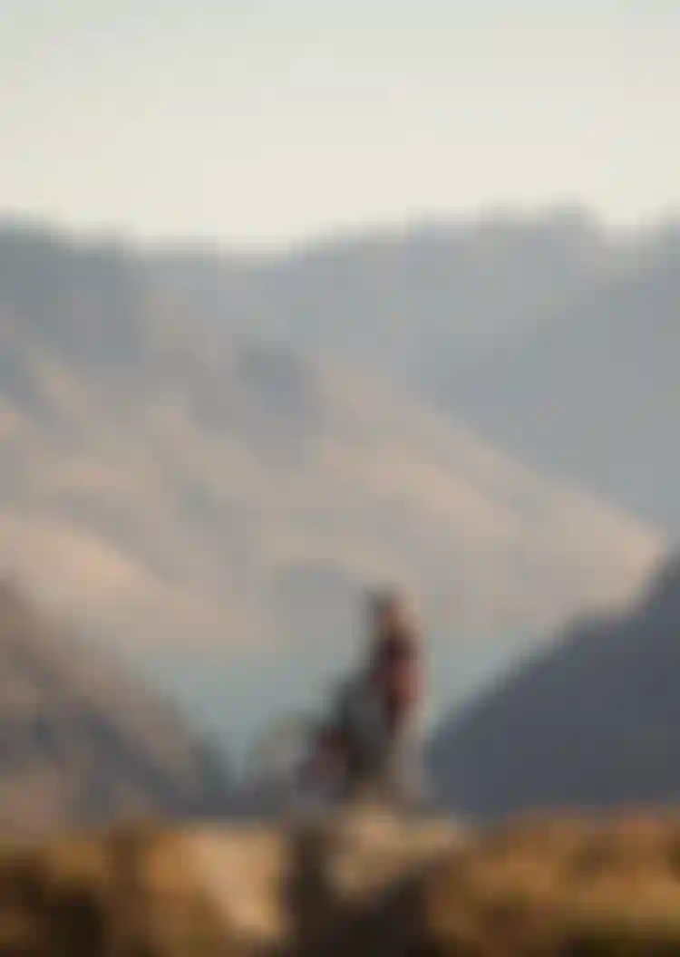Cyclist stands with mountain bike on a grassy hill, with rugged mountains and a lake in the background under a clear sky.