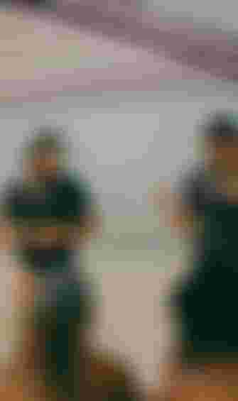 Two women wearing hats and black shirts stand under a canopy on a beach, speaking to seated people. The ocean is visible in the background.