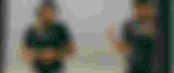 Two women wearing hats and black shirts stand under a canopy on a beach, speaking to seated people. The ocean is visible in the background.