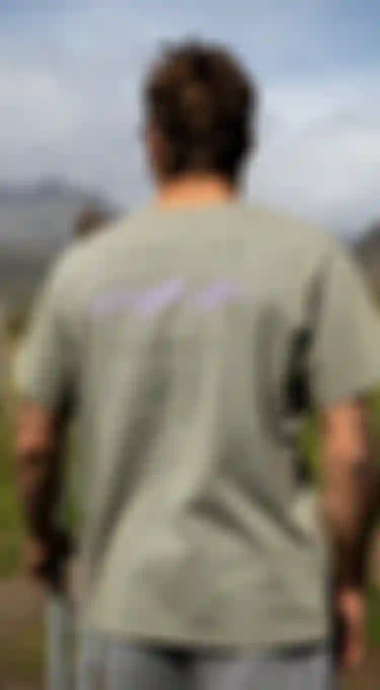 Backside of a man wearing a sage green tee with purple print standing on a green grass field