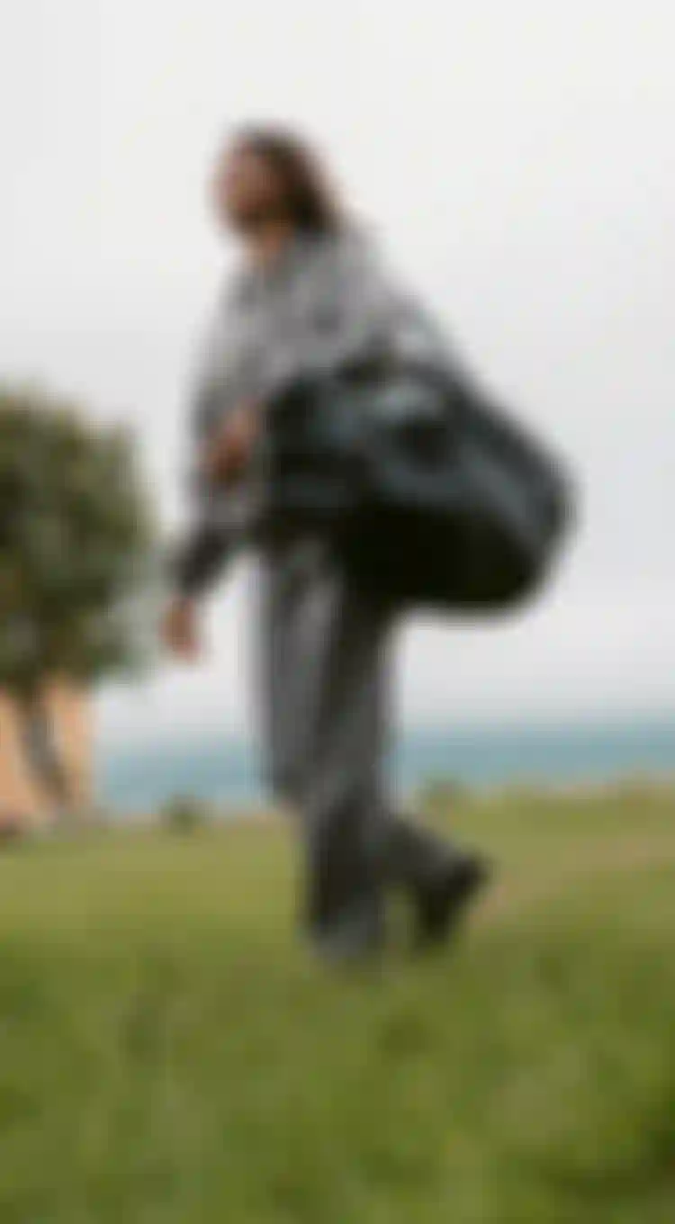 Woman in grey denim jacket and pants carrying a big duffle bag while walking on grass