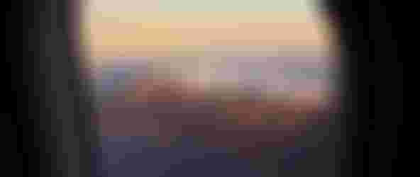 Airplane window framing a coastal mountain range topped by clouds, shoreline and deep blue ocean bathed in golden sunset light.