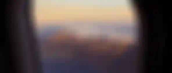 Airplane window framing a coastal mountain range topped by clouds, shoreline and deep blue ocean bathed in golden sunset light.
