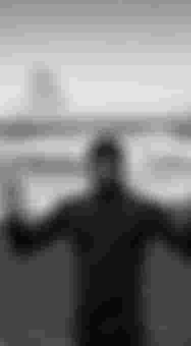 Man wearing a black wetsuit with hood standing on a beach with black sand, waves are breaking on the shore in the back ground. Rain is falling. Black and white image