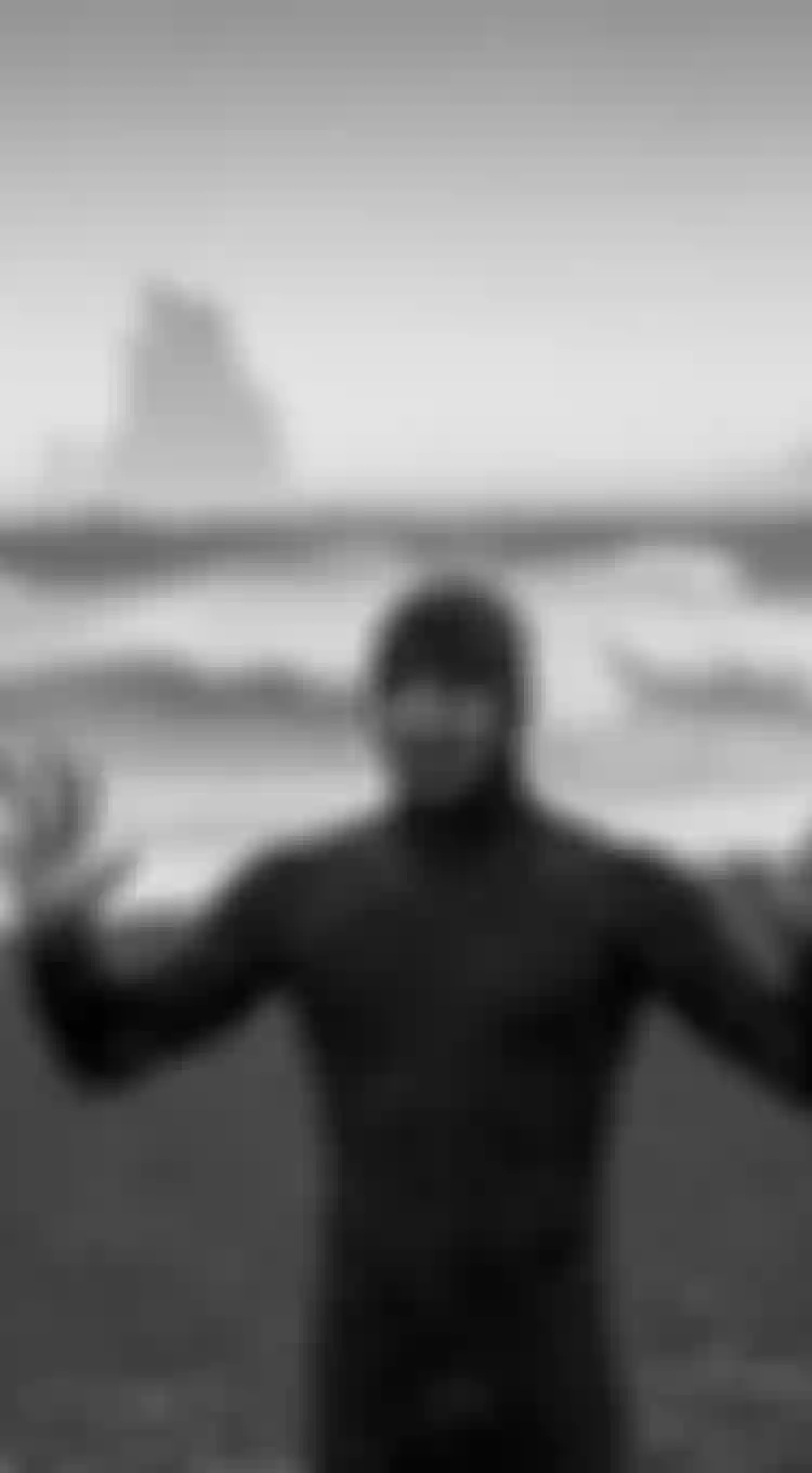 Man wearing a black wetsuit with hood standing on a beach with black sand, waves are breaking on the shore in the back ground. Rain is falling. Black and white image