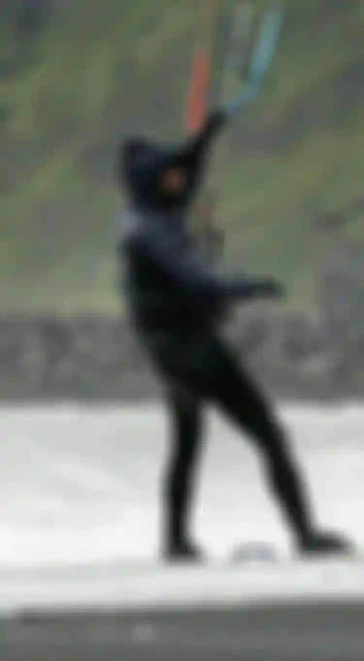 Man wearing a black wetsuit and neoprene jacket with hood on a kiteboard standing on the beach