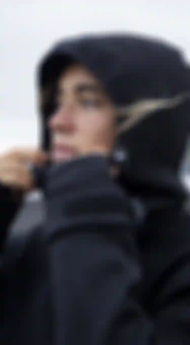 Close up of a woman wearing a black neoprene jacket with hood, blurred beach with black sand in the background