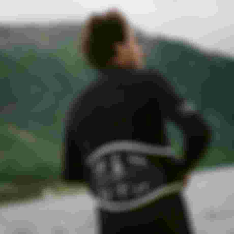 Man wearing a black wetsuit and kite harness in front of green mountains