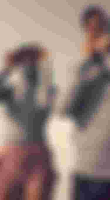 Two people on a sandy beach wearing sun-protective clothing and hats, adjusting their masks, with a cloudy sky in the background.