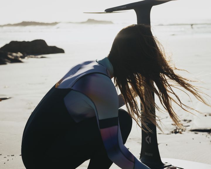 Woman in a wetsuit adjusts a foil board on a sandy beach, with long hair blowing in the wind and a scenic ocean backdrop.