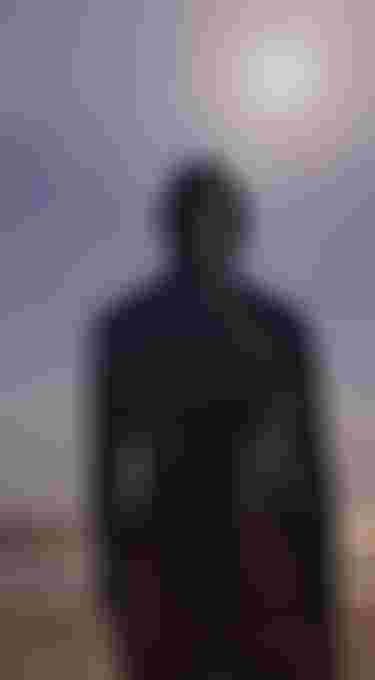 Person with a hood stands on a beach, facing the ocean, with the sun shining brightly in the sky.