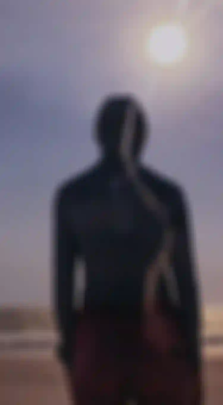 Person with a hood stands on a beach, facing the ocean, with the sun shining brightly in the sky.