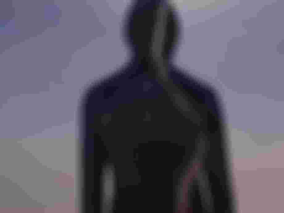 Person with a hood stands on a beach, facing the ocean, with the sun shining brightly in the sky.