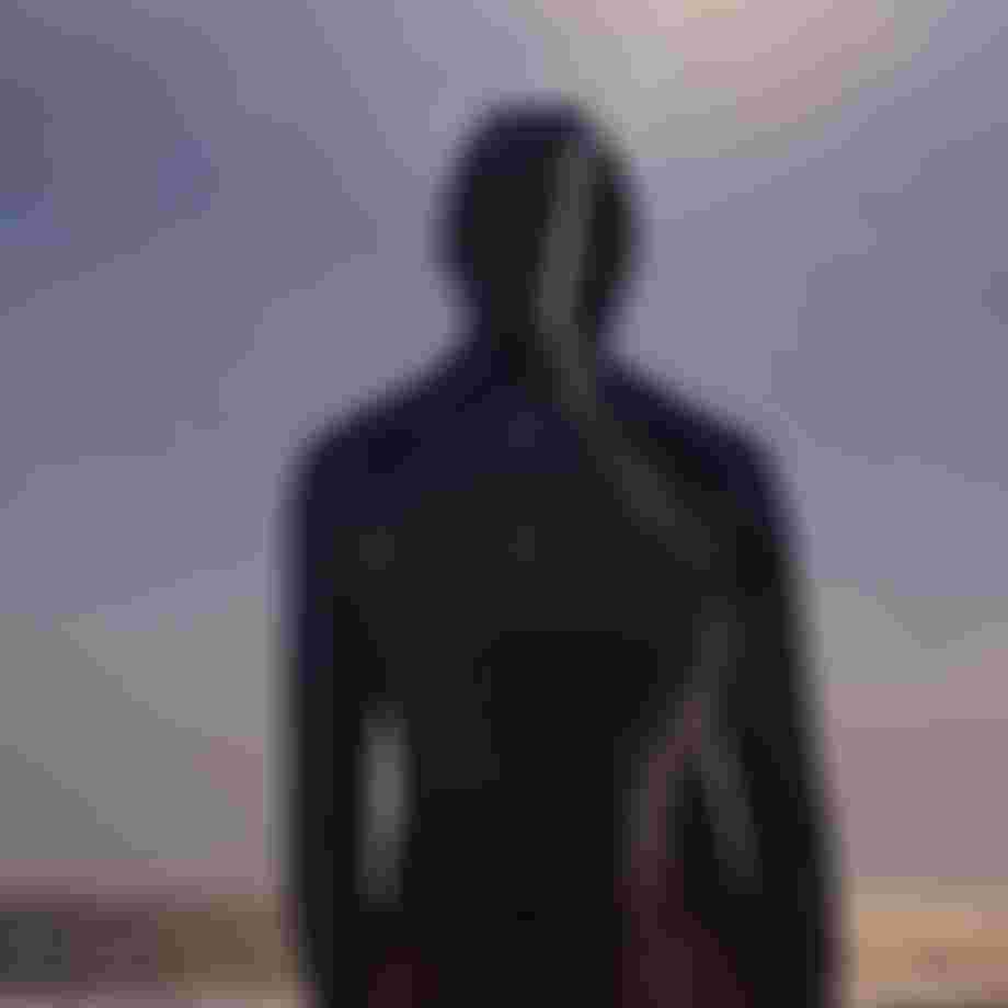 Person with a hood stands on a beach, facing the ocean, with the sun shining brightly in the sky.