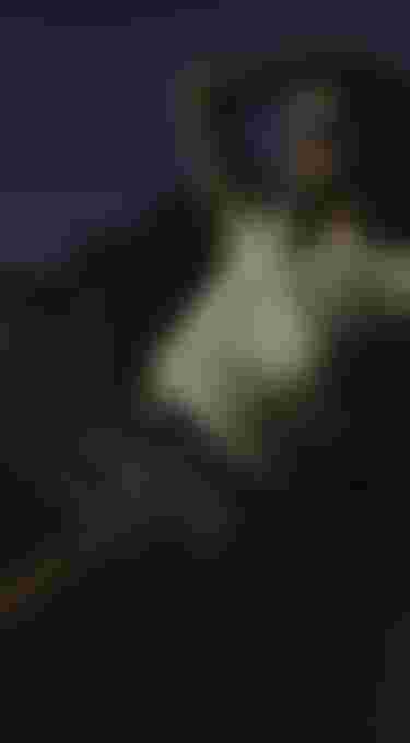 Woman in a yellow t-shirt and blue shorts leaning against rocks, looking into the sun with sunglasses on